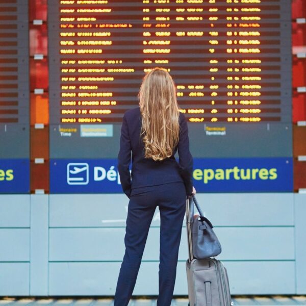 image of woman with light brown hair looking at a flight tracker board in the airport. the woman is wearing a black pantsuit and black heels, and is holding luggage