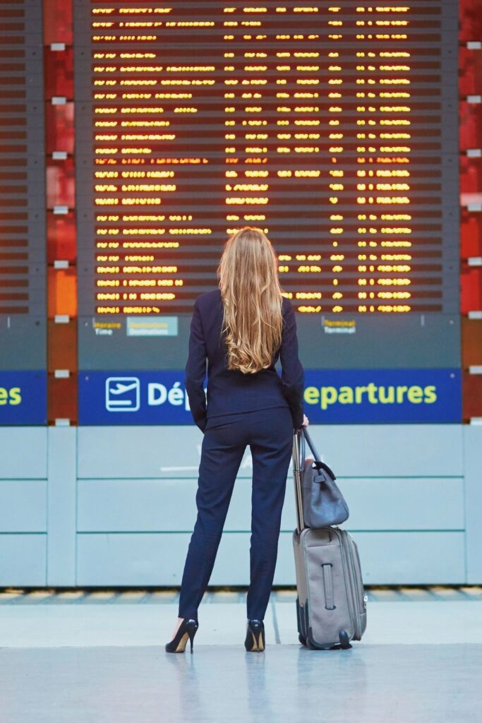 image of woman with light brown hair looking at a flight tracker board in the airport. the woman is wearing a black pantsuit and black heels, and is holding luggage