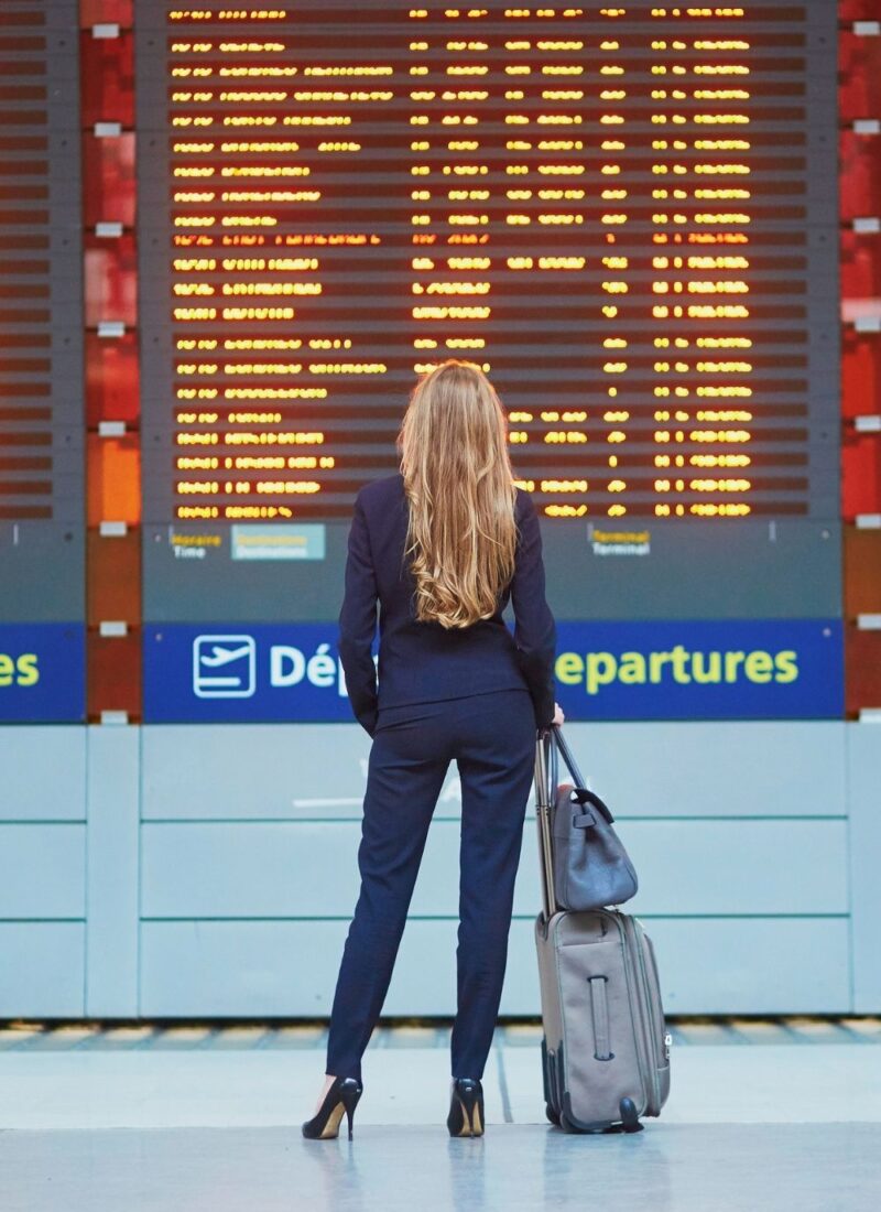 image of woman with light brown hair looking at a flight tracker board in the airport. the woman is wearing a black pantsuit and black heels, and is holding luggage