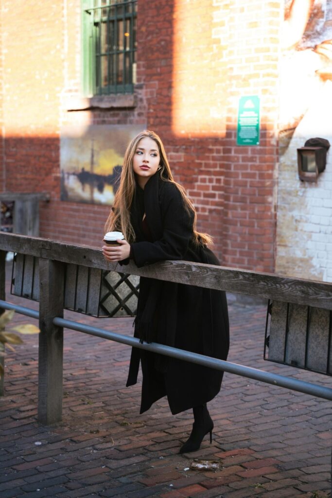 Image of woman after checking into Airbnb, with brown hair in black trenchcoat leaning over railing beside a street, coffee in hand