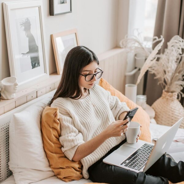 Image of woman with white sweater and black glasses sitting in bed with laptop on lap, holding a phone