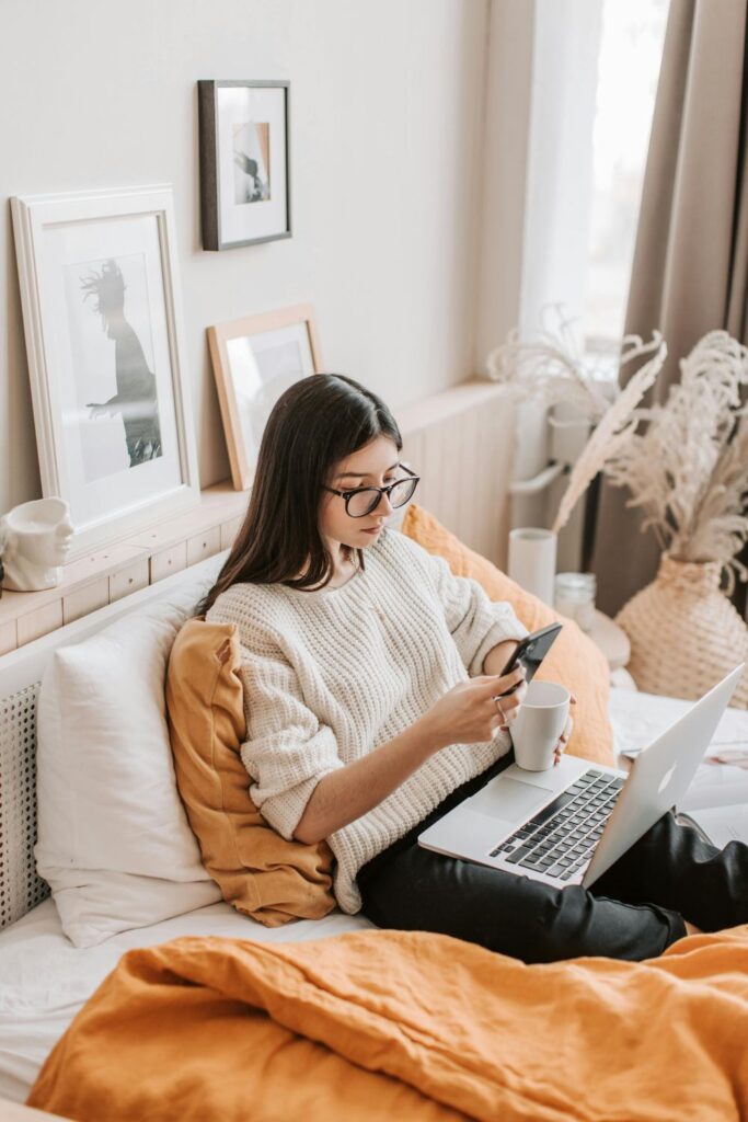 Image of woman with white sweater and black glasses sitting in bed with laptop on lap, holding a phone