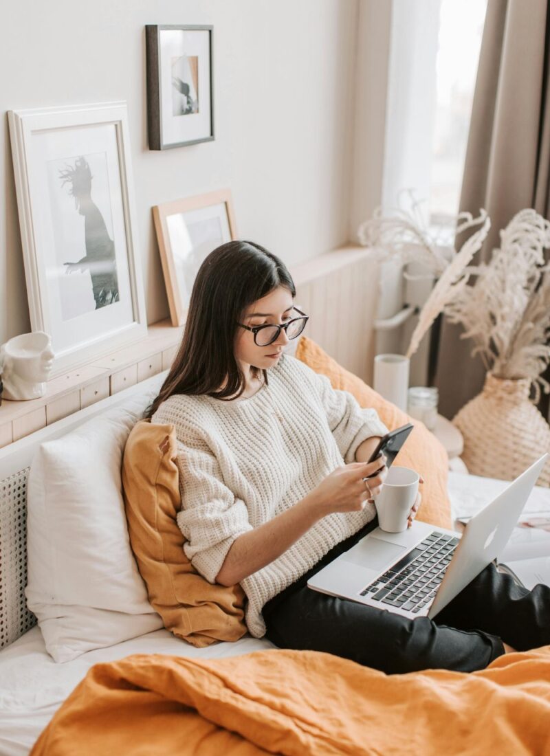 Image of woman with white sweater and black glasses sitting in bed with laptop on lap, holding a phone