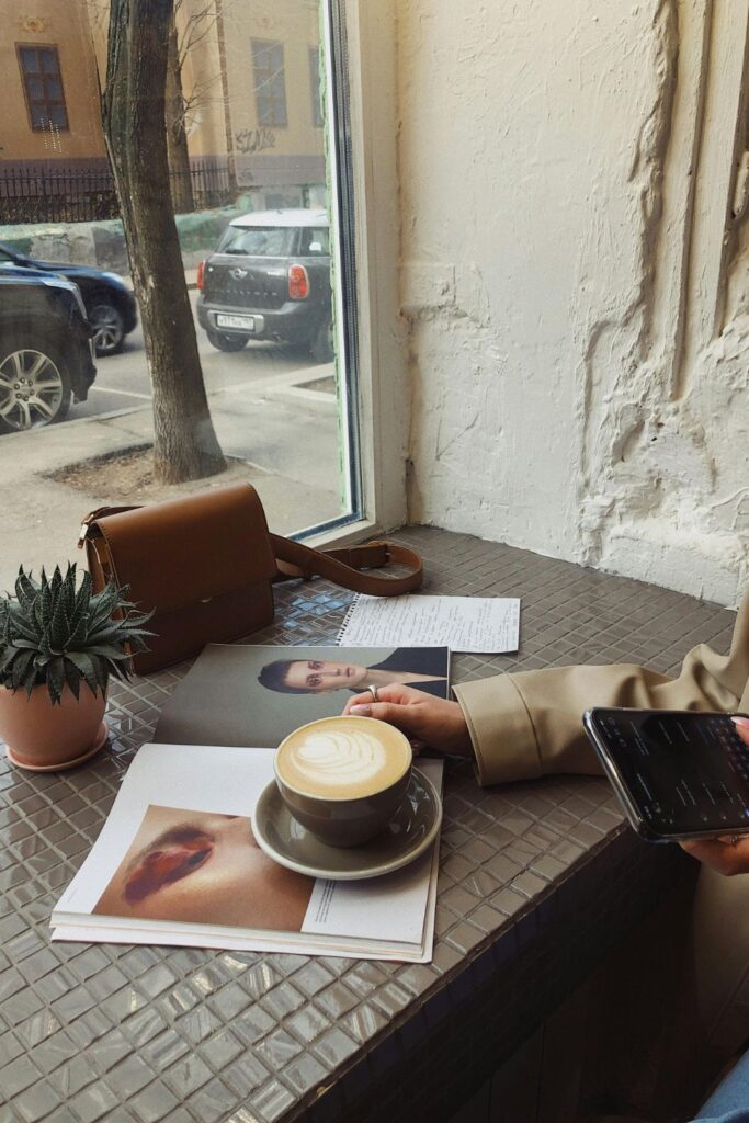 Image of cafe table with magazine and coffee mug with latte set on table. The table overlooks a window and the street