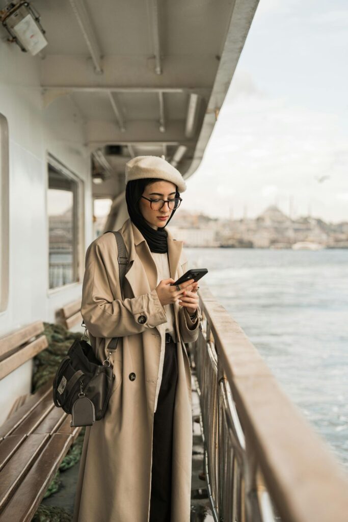 Image of woman in tan trench coat, black scarf and cream hat, standing on the side of a passenger ship overlooking the water. Woman is holding a black smart phone and looking at the screen to speak to Marco Polo app users