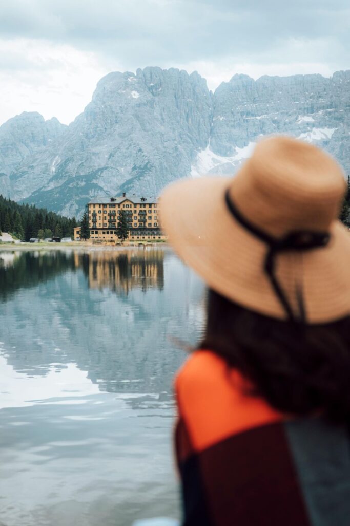 Image of woman facing away from camera, wearing black and orange sweater and straw hat. Woman is facing a view of a lake and mountain