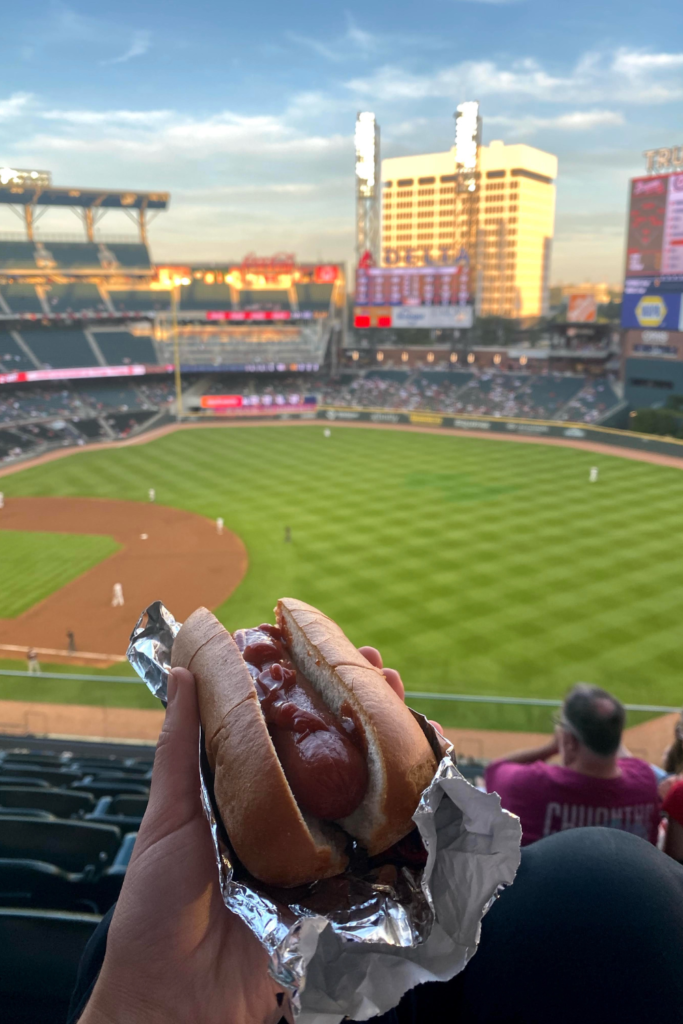 Image of hand holding up a hot dog in tin foil with a baseball stadium in the background 