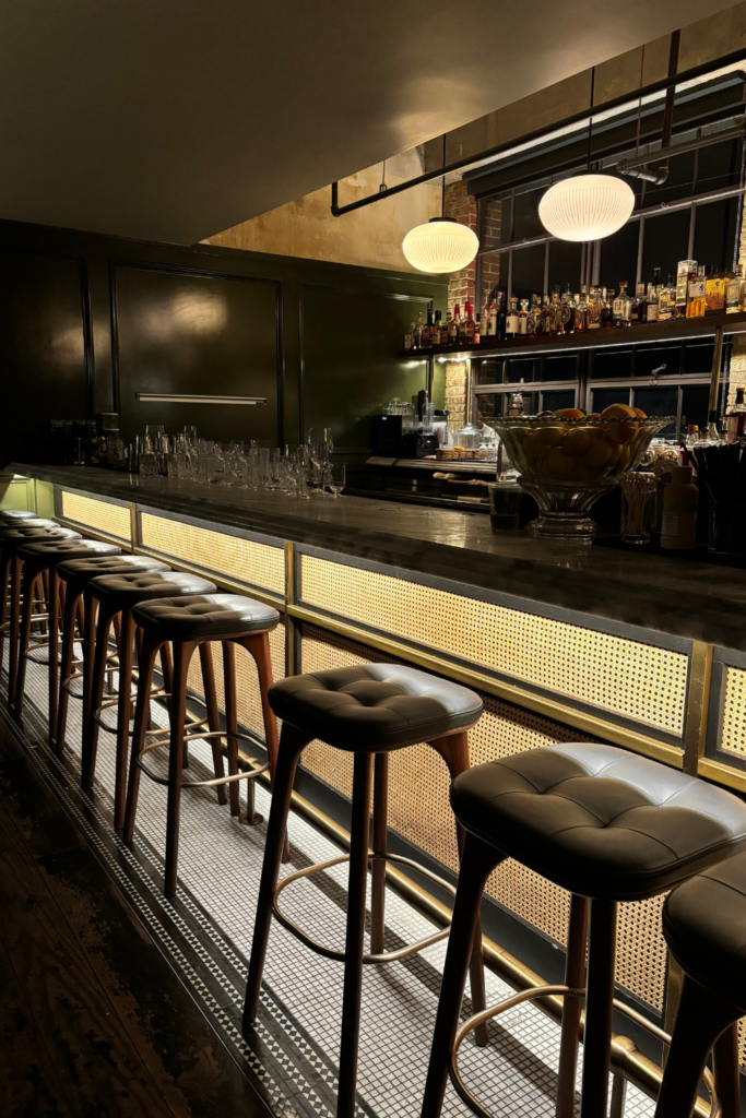 A view of the dimly lit bar top and stools at 12 Cocktail Bar in the top of Ponce Market, an excellent date spot for couples in Atlanta Georgia. The image shows the stools in foreground with a dim overhead lighting and various bottles of alcohol on a shelf against the wall.