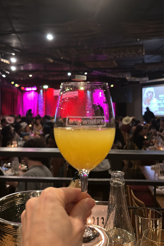 First person point of view of holding up a mimosa in a glass in front of a stage lit with pink and red lights at City Winery in Atlanta Georgia