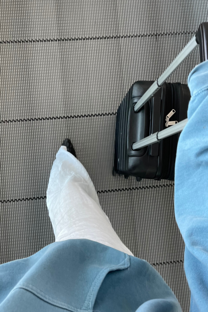 First person point of view looking down at someone's shoes walking through an airport. The person in the image is wearing a blue top and white pants, and wheeling a suitcase in the right hand.