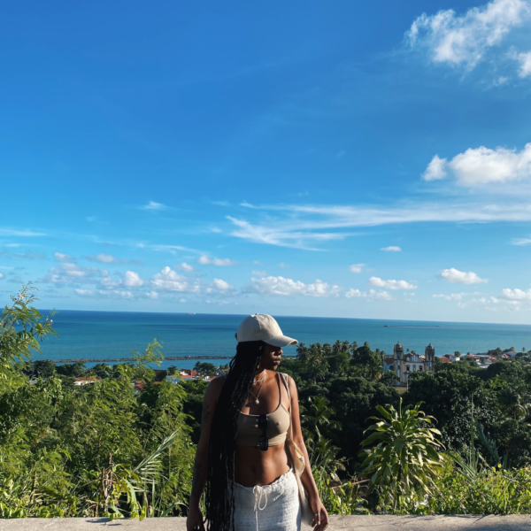 Image of woman in foreground of photo looking to the side, with the ocean and greenery in the background on a sunny day.