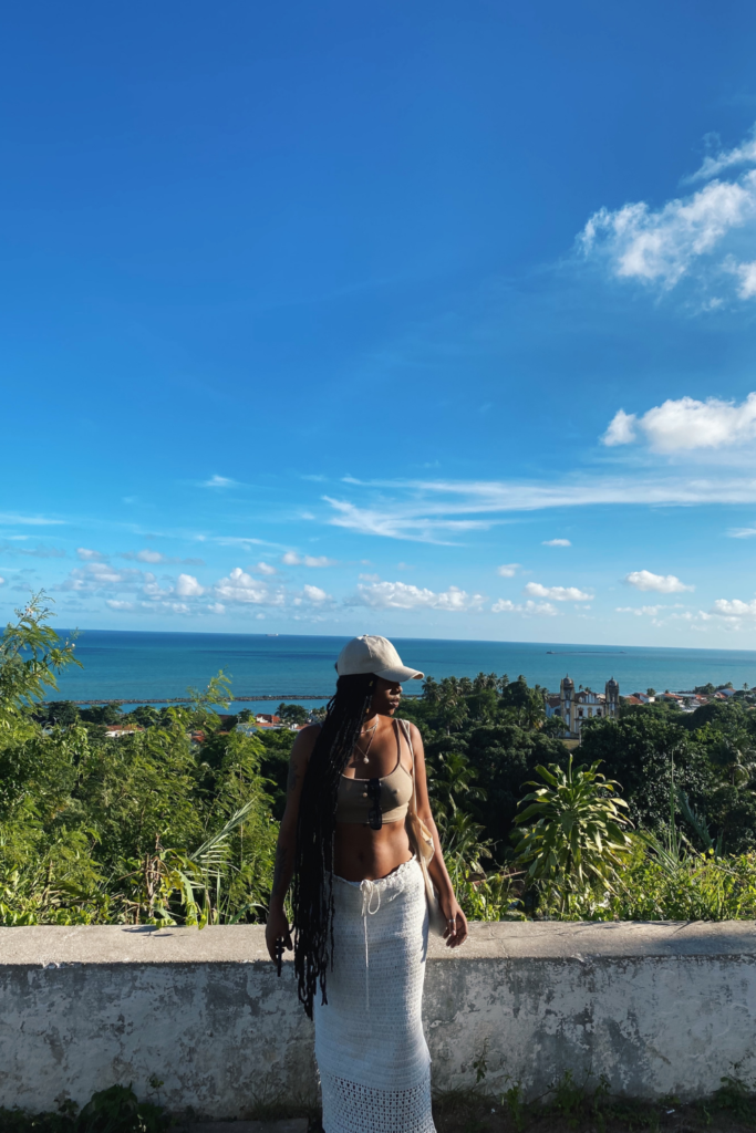 Image of woman in foreground of photo looking to the side, with the ocean and greenery in the background on a sunny day.