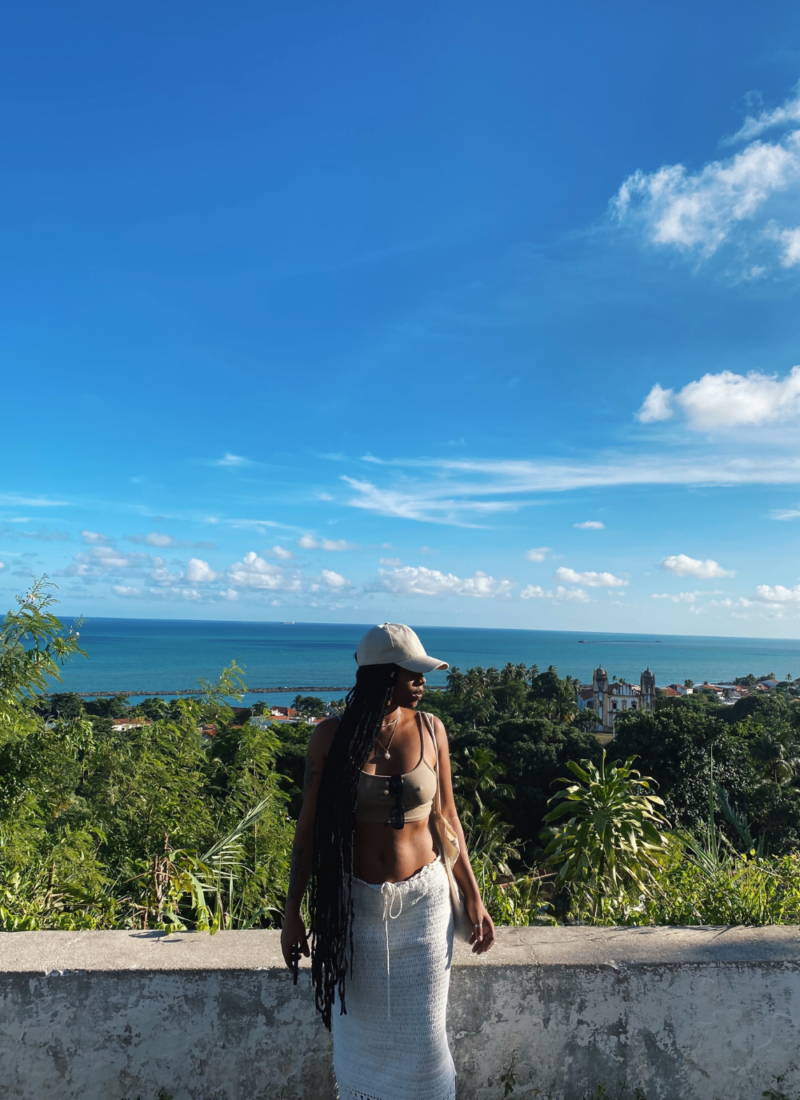 Image of woman in foreground of photo looking to the side, with the ocean and greenery in the background on a sunny day.