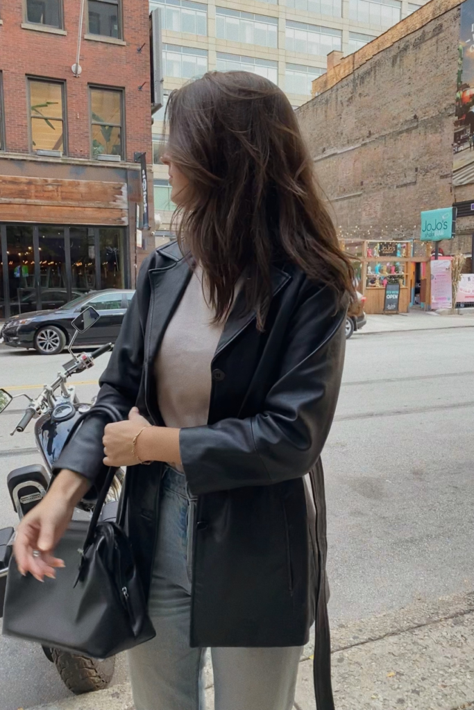 Image of woman in leather jacket standing in city looking away from camera holding small bag used for carry on during travels