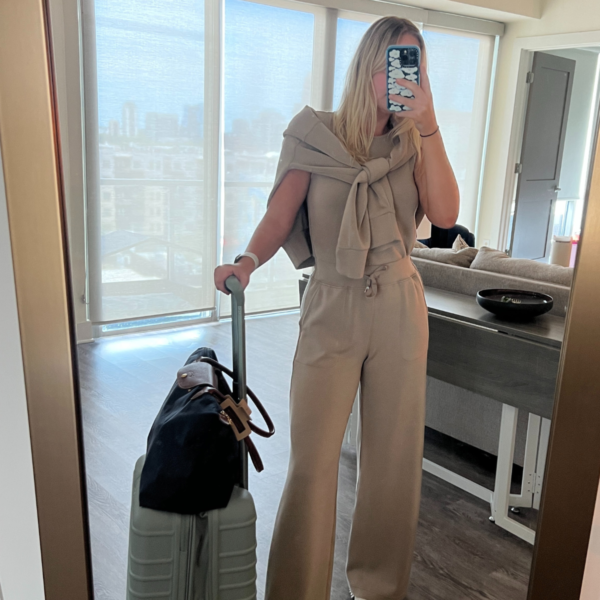 Image of woman in mirror wearing tan travel outfit with carry on luggage that is one inch too big