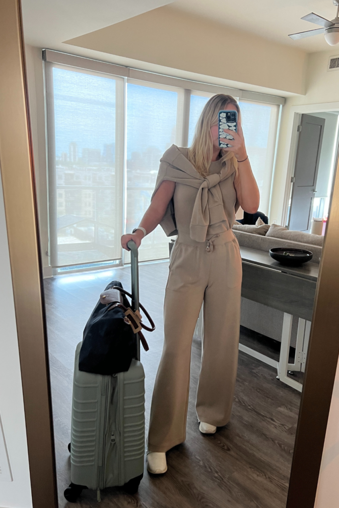 Image of woman in mirror wearing tan travel outfit with carry on luggage that is one inch too big