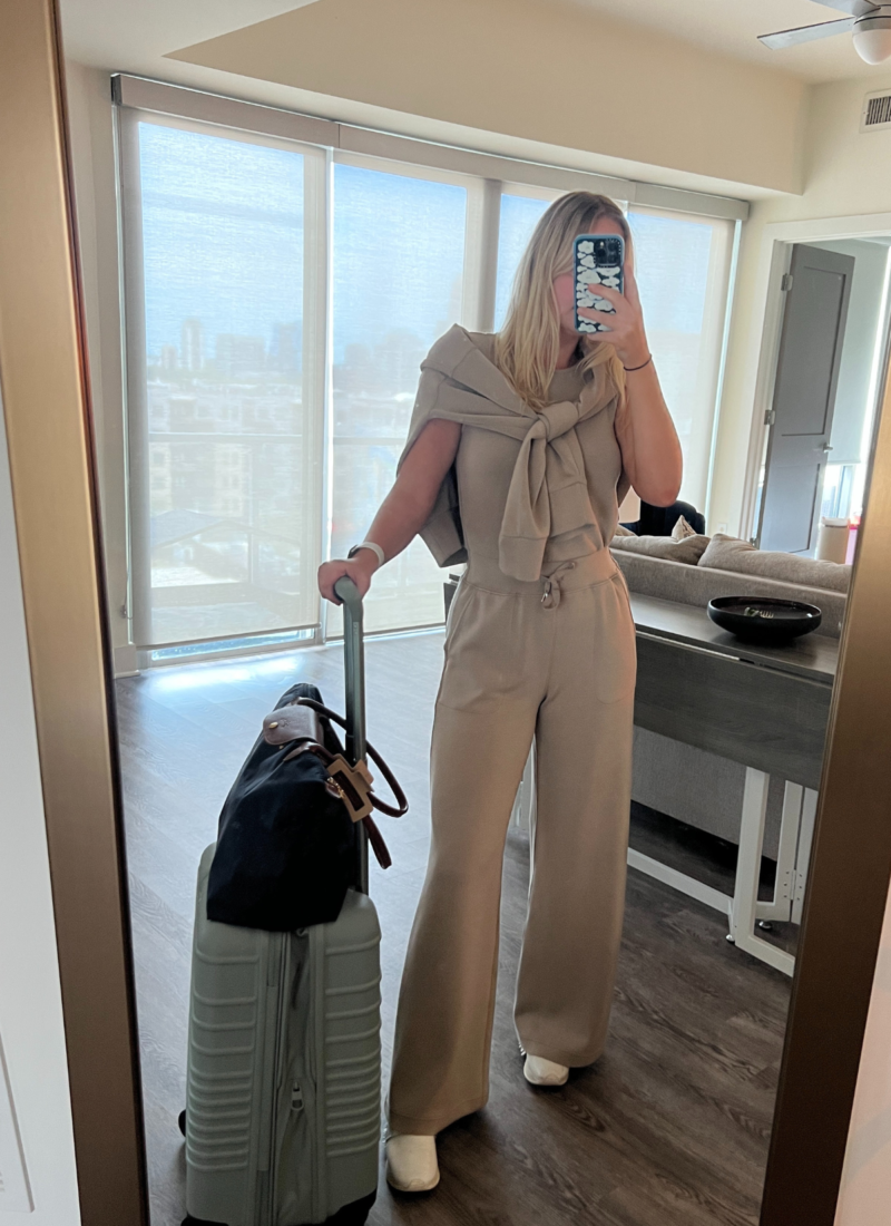 Image of woman in mirror wearing tan travel outfit with carry on luggage that is one inch too big