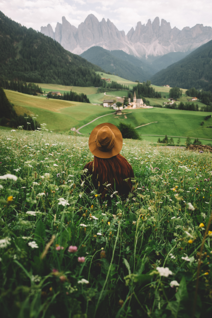 Image of woman in hat sitting in field of flowers, facing away from camera and looking at mountains in distance.