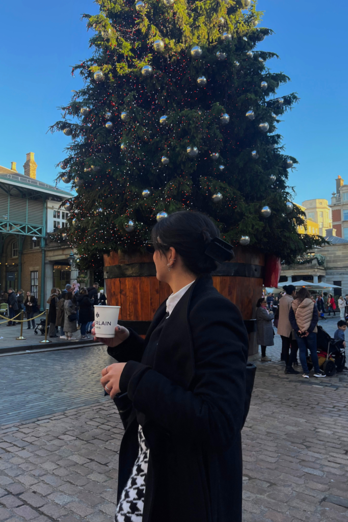 Image of woman holding coffee cup and looking towards a Christmas tree outdoors.