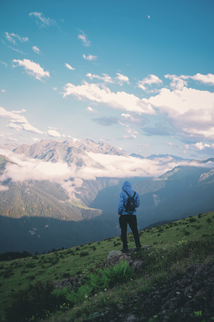 Image of traveler standing on a tall hilltop, facing away from camera and looking at tall mountains in distance.