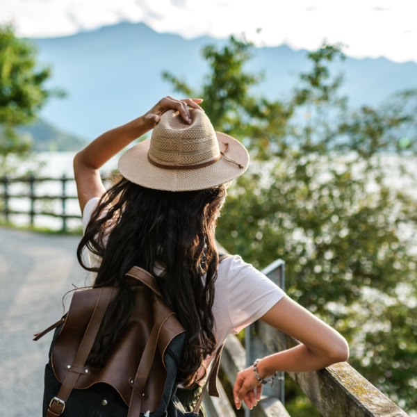 image of woman facing away from camera looking at a mountain and lake scene. Woman is wearing a white hat and backpack.