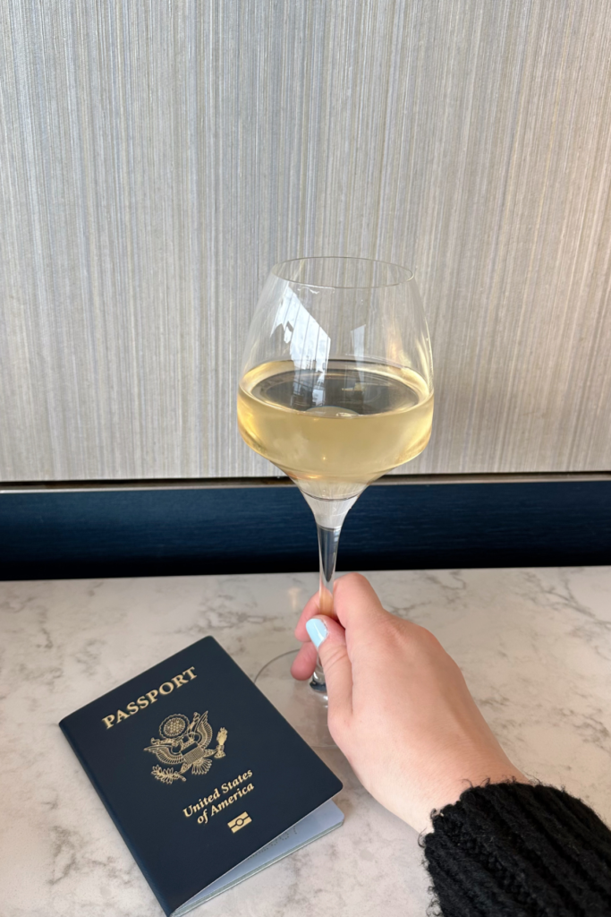 Hold holding a glass of white wine, with a passport resting on the table beside the wine.