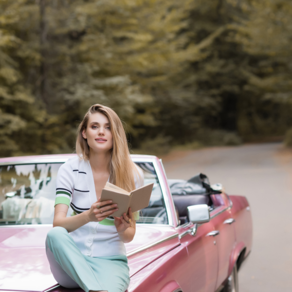 Image of a woman holding a book open sitting on the hood of a red car on the side of the road with trees in background.