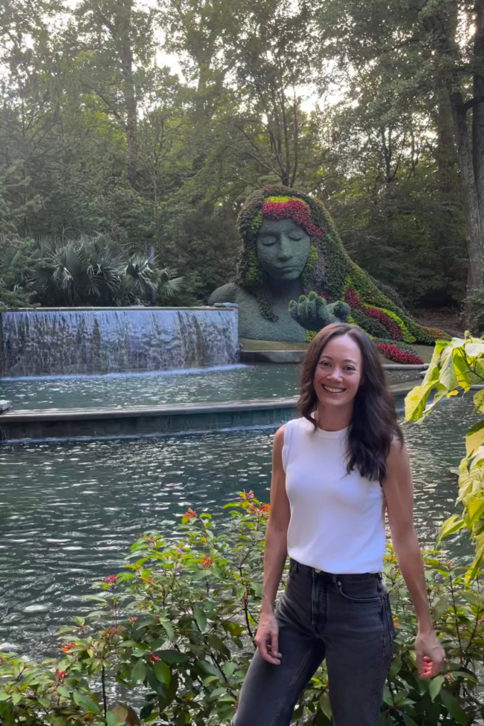 Image of woman with brown hair and white top standing in front of water fall at Atlanta Botanical Garden.